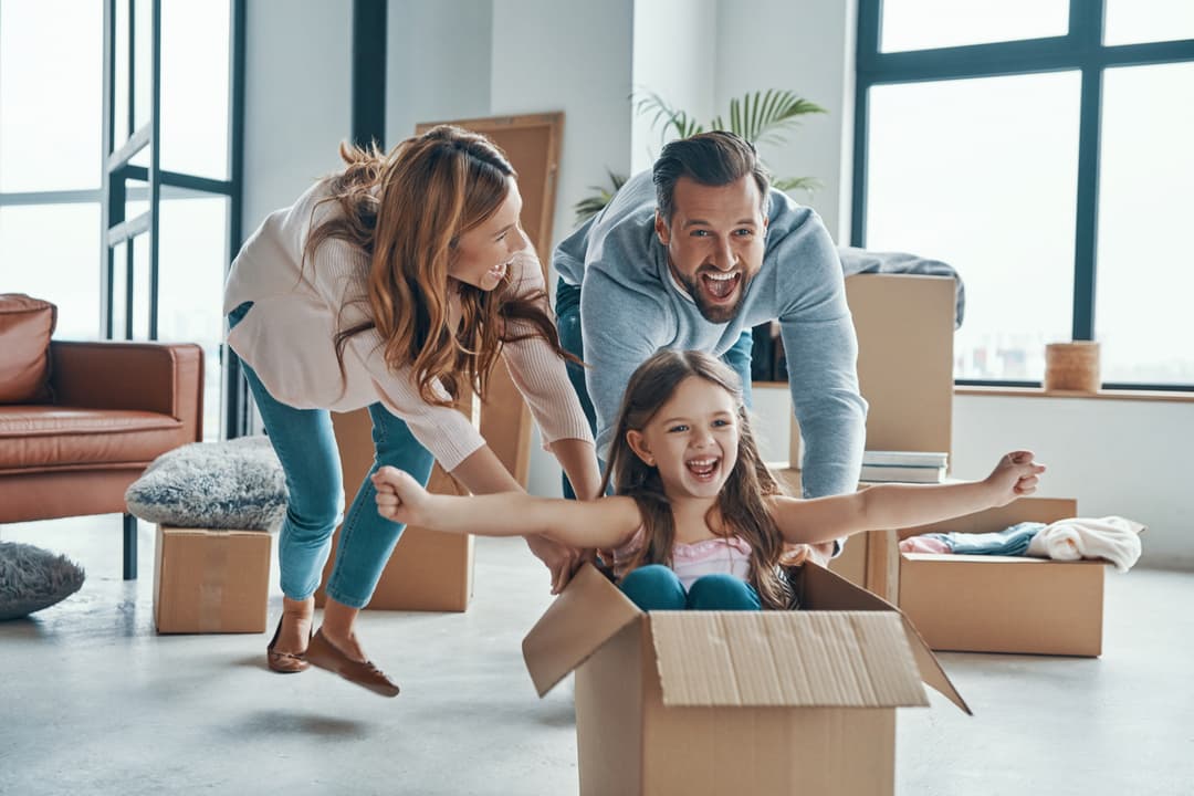 Mutter und Vater schieben Kind in einer Umzugskiste durch das Haus